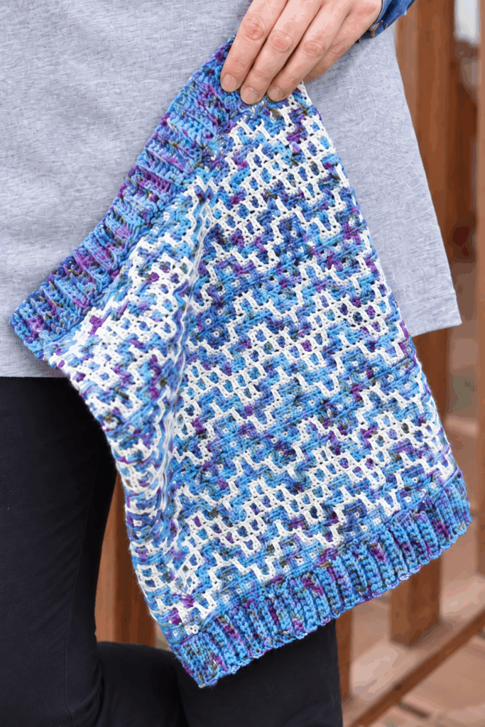 Drutozlot mosaic crochet cowl in variegated blue and purple with white, held in one hand to show the mosaic pattern and ribbed edges
