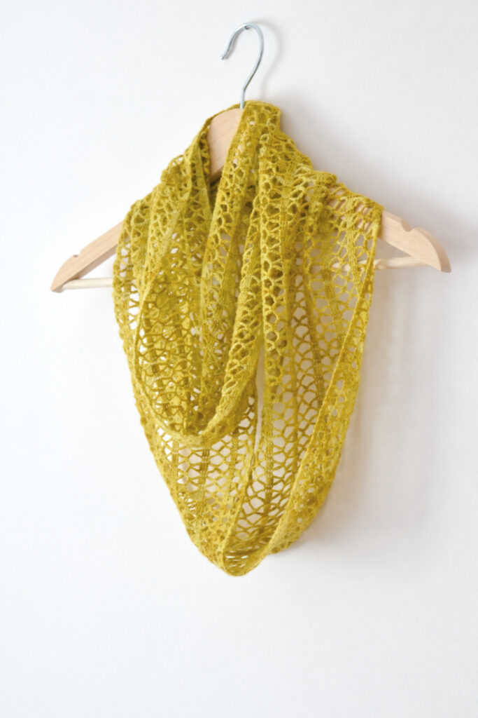 Crochet lace cowl in ochre draped on a wooden hanger against a white wall