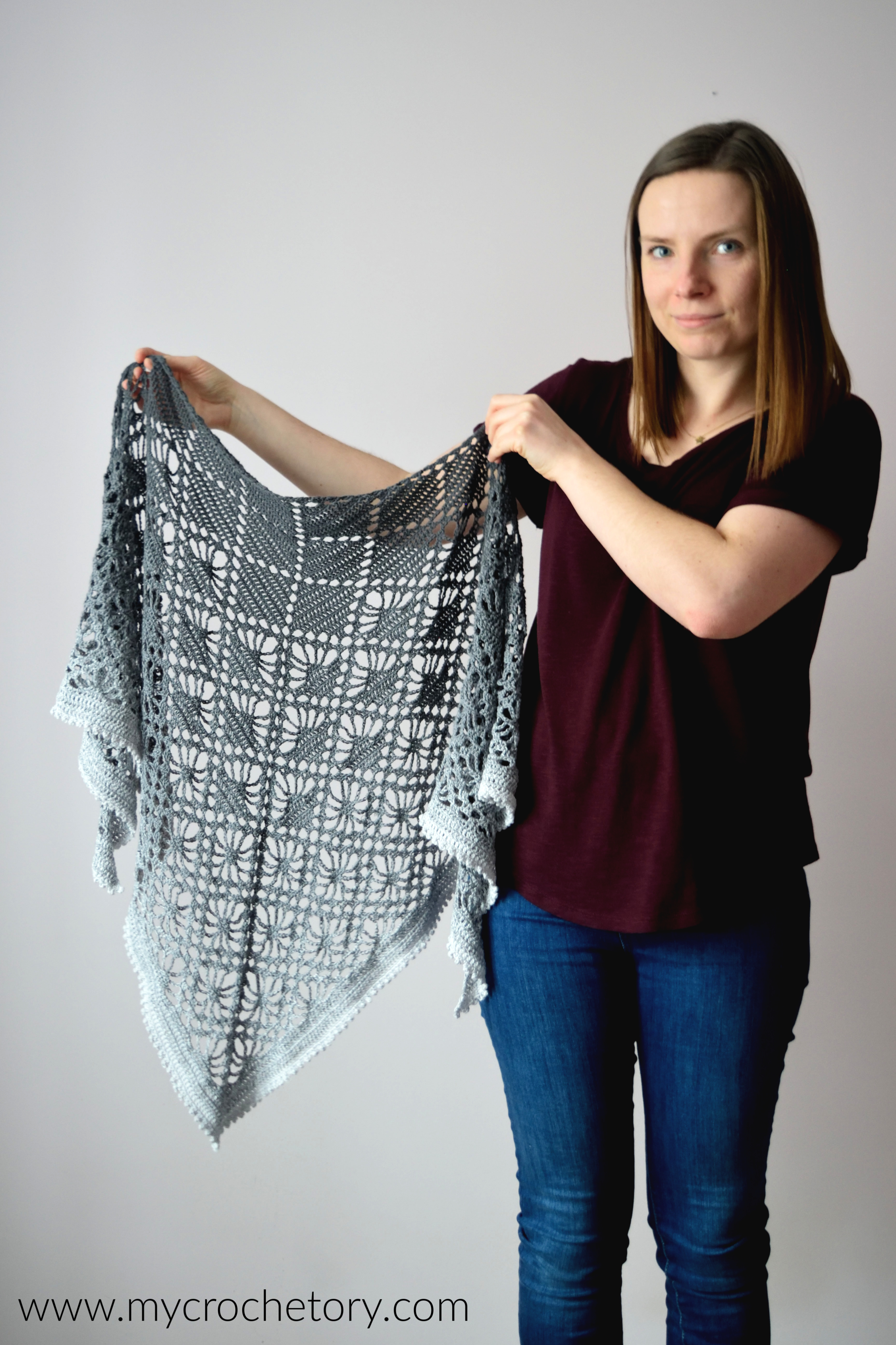 Woman holding up a finished diamond lace crochet shawl in a gray gradient