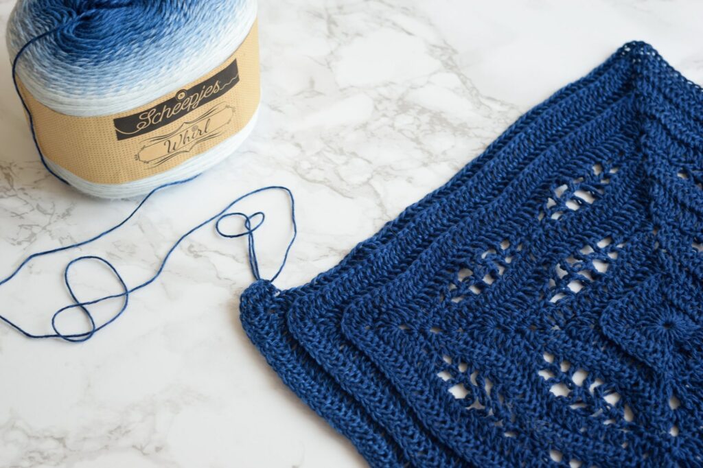 Kalinda Blanket in progress in navy blue, next to a cake of Scheepjes Whirl yarn on a marble table