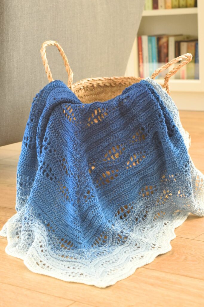Kalinda crochet blanket in blue and white ombre, hanging out of a woven basket with handles