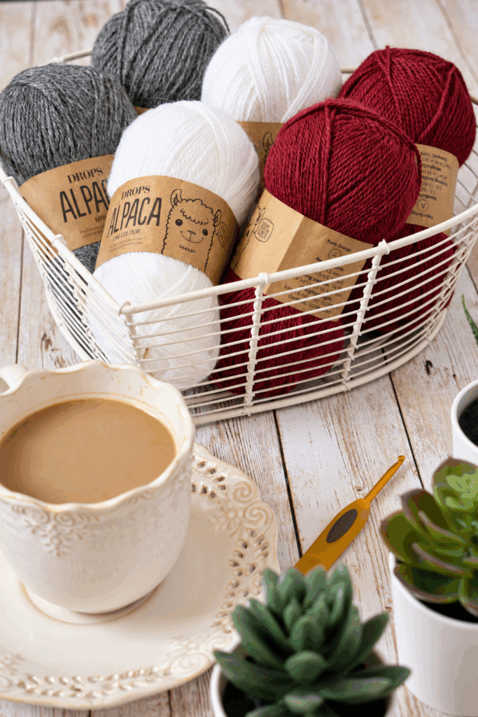 	DROPS Alpaca yarn in white, gray and red in a wire basket, with a crochet hook and a cup of coffee