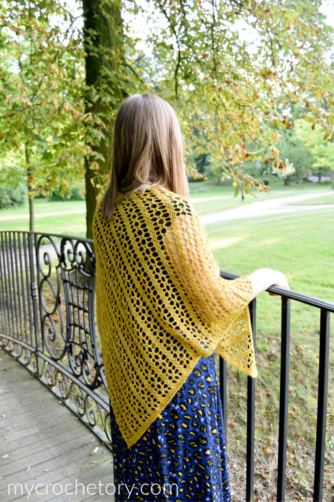 Lace triangle shawl in mustard gold draped over the shoulders with the point at the back, by an iron railing