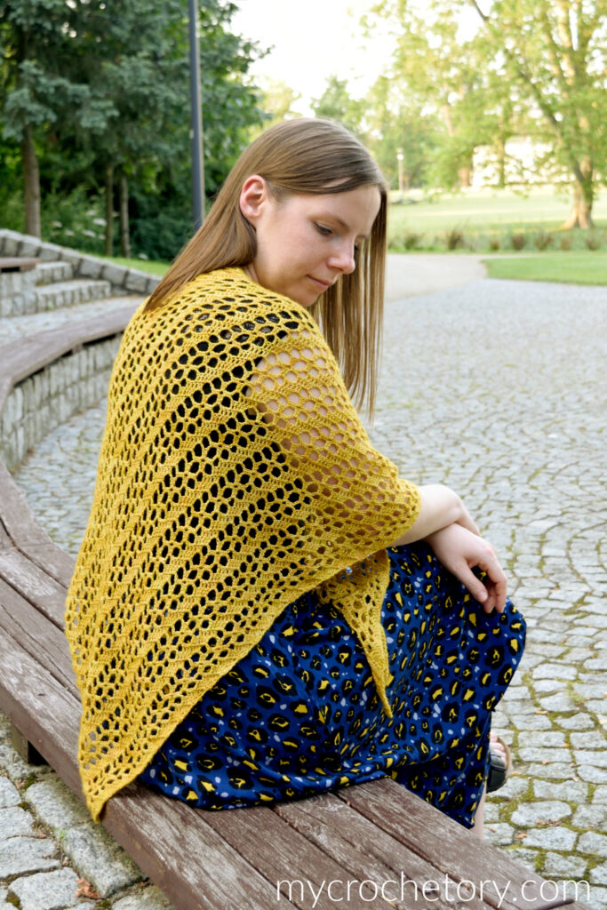 Woman sitting on a curved wooden bench wearing a mustard crochet shawl over her shoulders