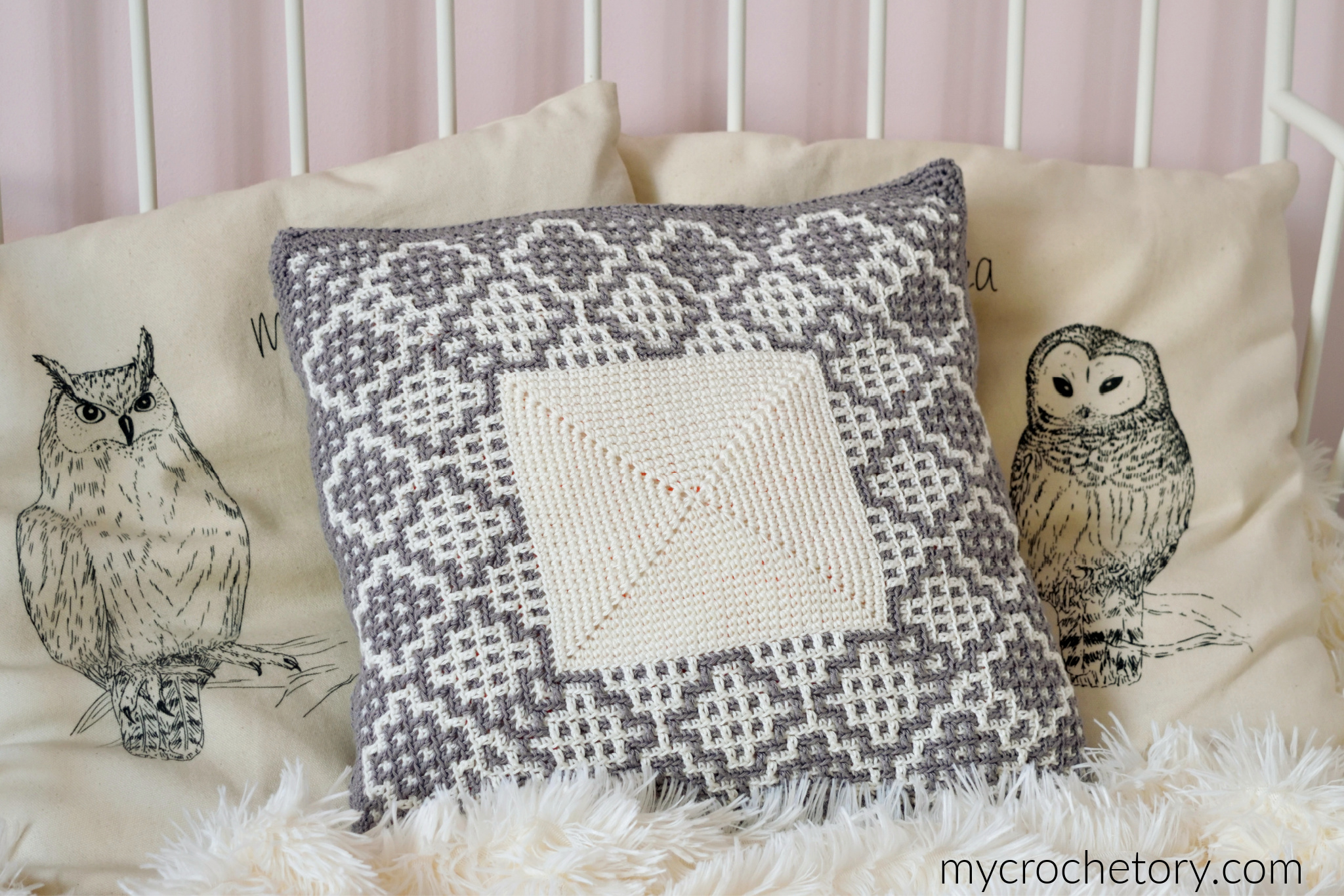 Crochet square pillow with a mosaic border between two owl print pillows on a bed