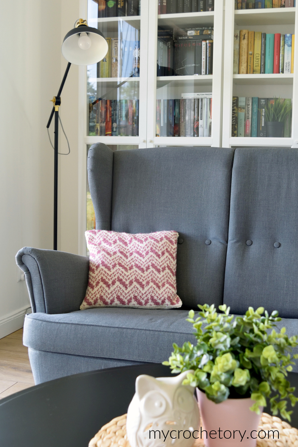 Cevro intarsia crochet pillow on a gray sofa in a living room with a bookcase and a lamp