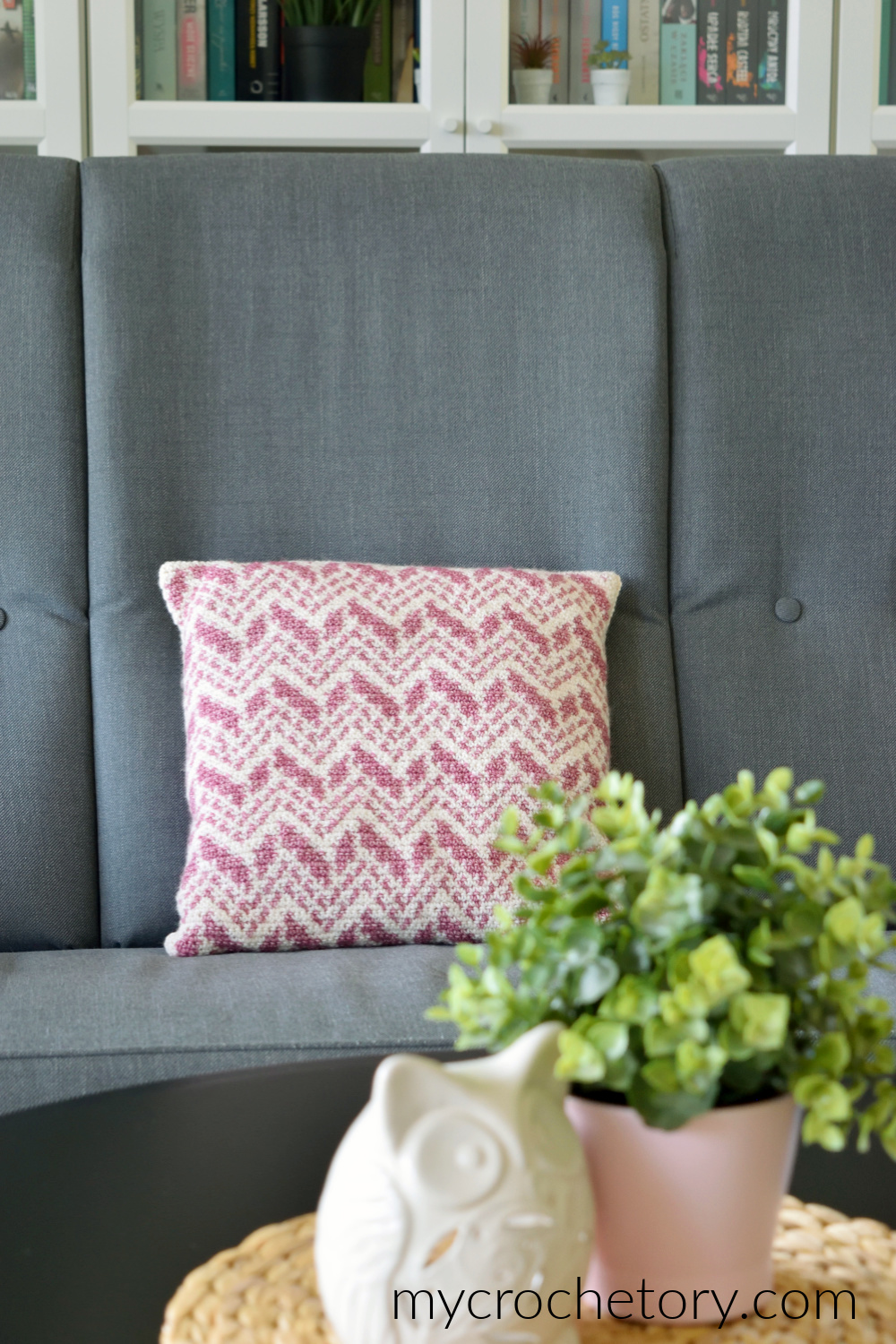 Intarsia crochet pillow with cream and pink chevrons on a gray armchair next to a plant
