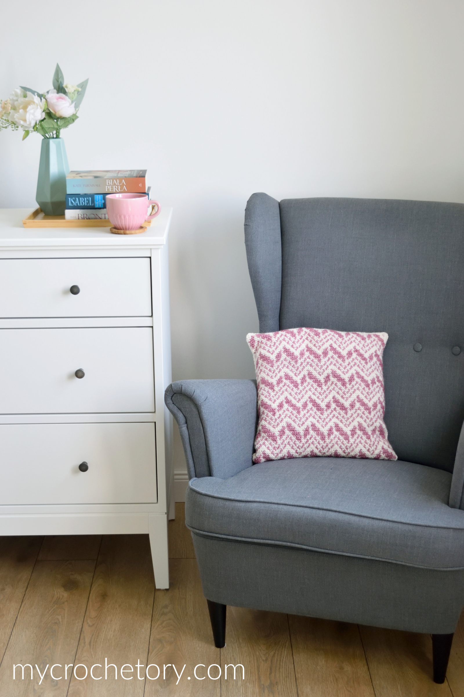 Intarsia crochet pillow on a gray armchair next to a white dresser with flowers