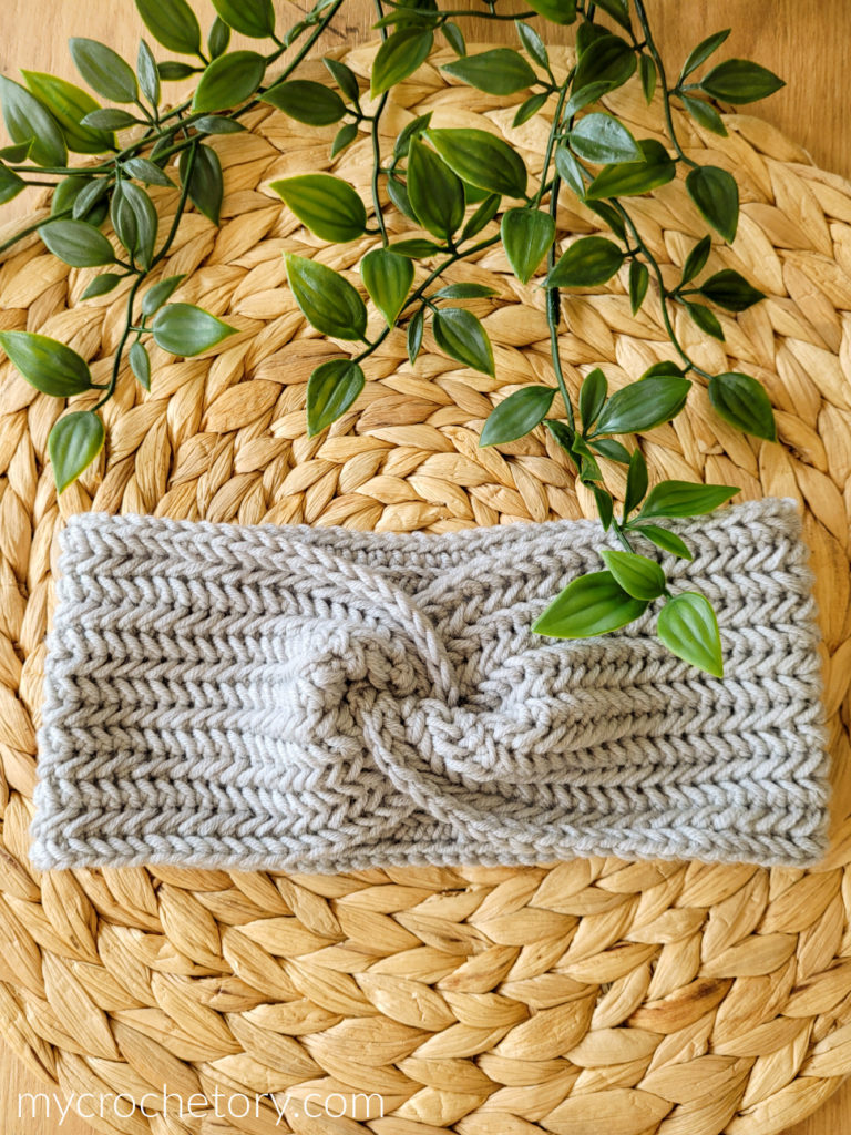 Twisted herringbone crochet headband in light gray on a woven placemat with green leaves