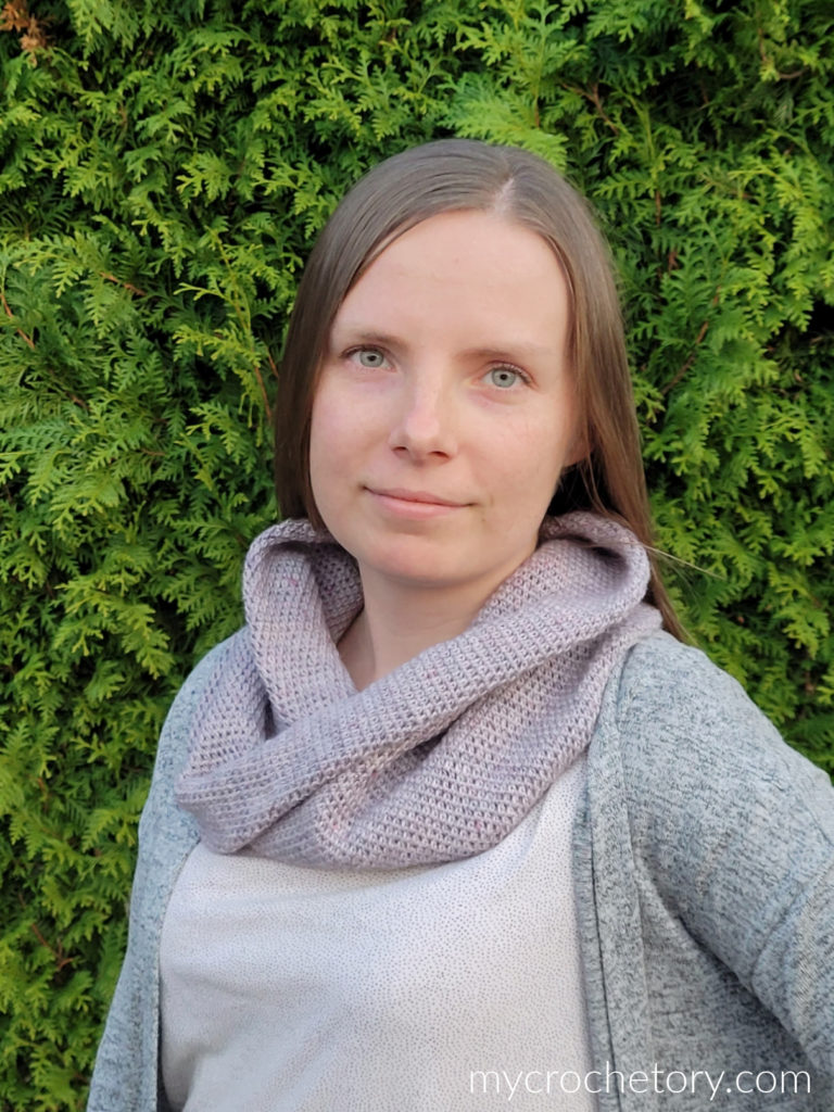 Lilac Moebius crochet cowl in pale lilac with a knit-look texture, worn with a gray cardigan in front of a green hedge