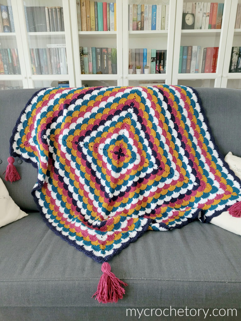 Clamshell Square Blanket in pink, teal, mustard and white, worked from the center out, with pink tassels on a gray sofa