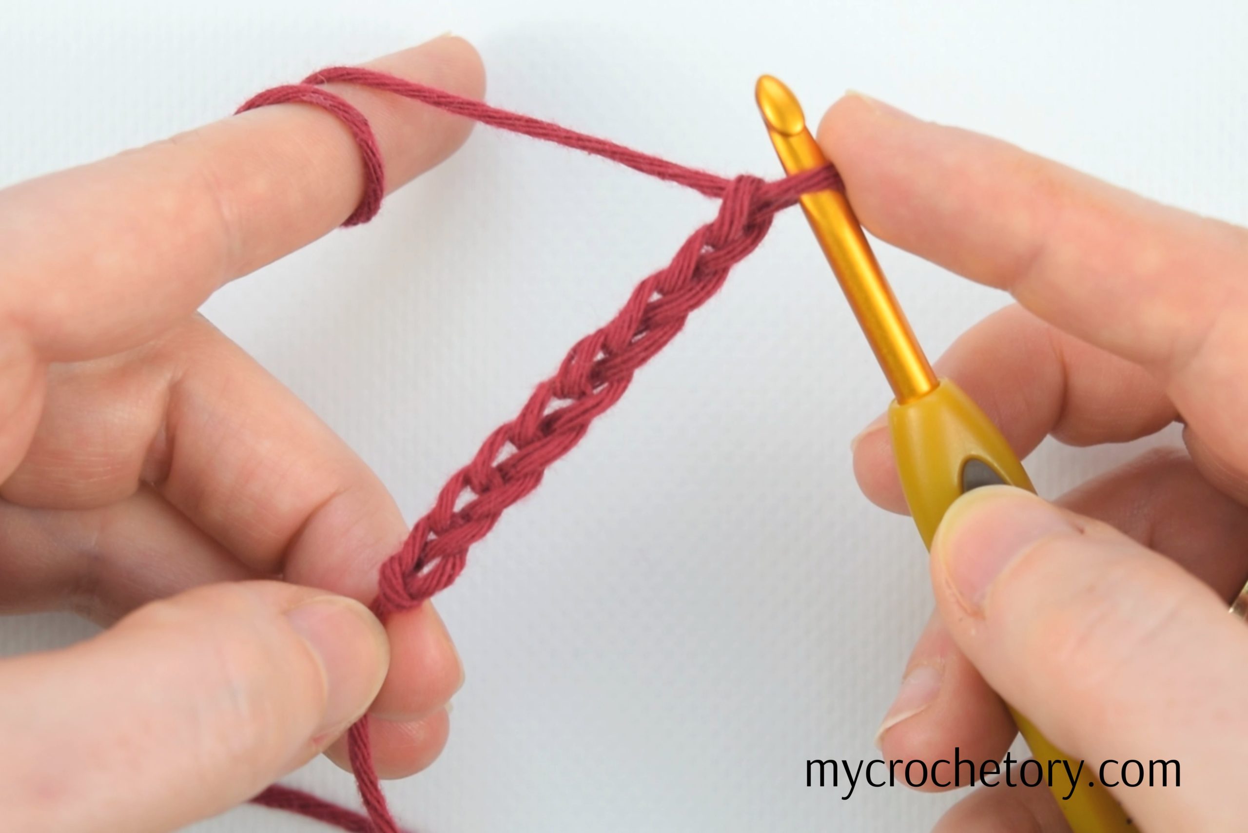Hands making a foundation chain with red yarn and a gold crochet hook