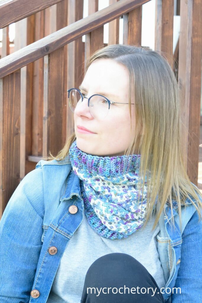 Woman wearing a colorful mosaic crochet cowl pattern with ribbed edging, sitting outdoors in a denim jacket