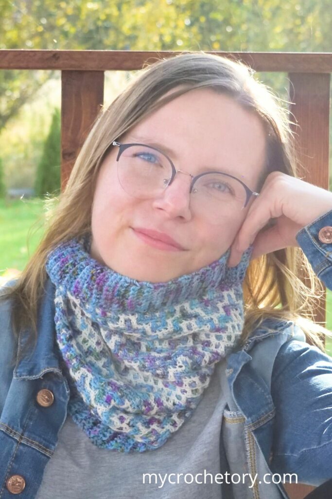 Close-up of a woman smiling while wearing a blue and purple mosaic crochet cowl pattern against a natural green background