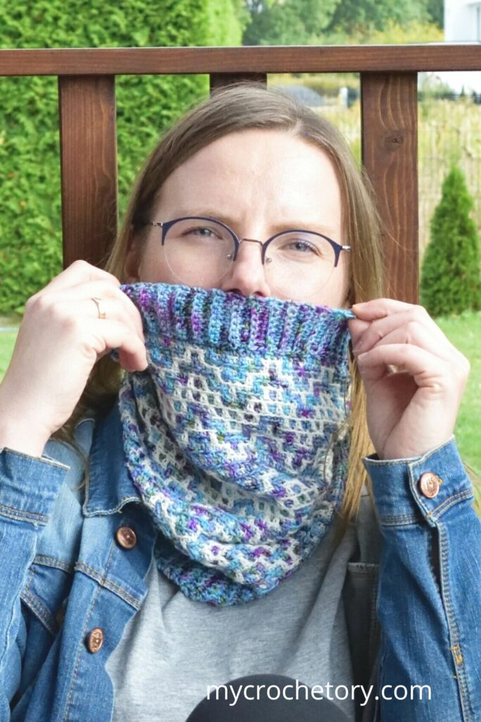 Woman playfully holding up her mosaic crochet cowl pattern, covering part of her face, showcasing the detailed design.