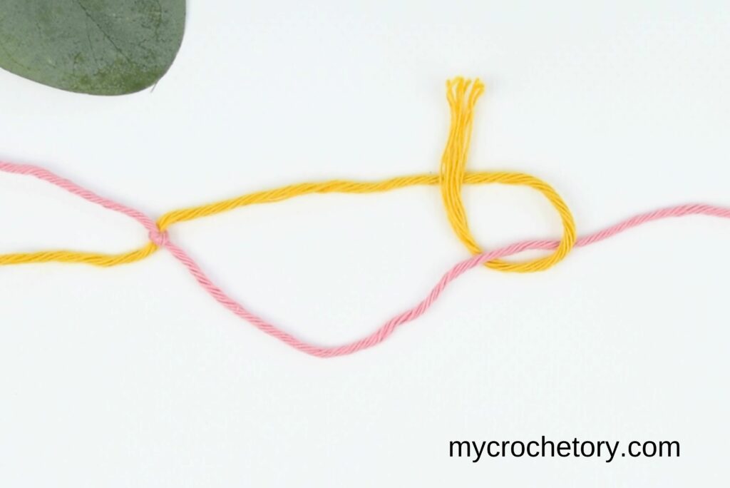Yellow yarn looped over itself after going under the pink yarn, completing the magic knot for joining yarn.