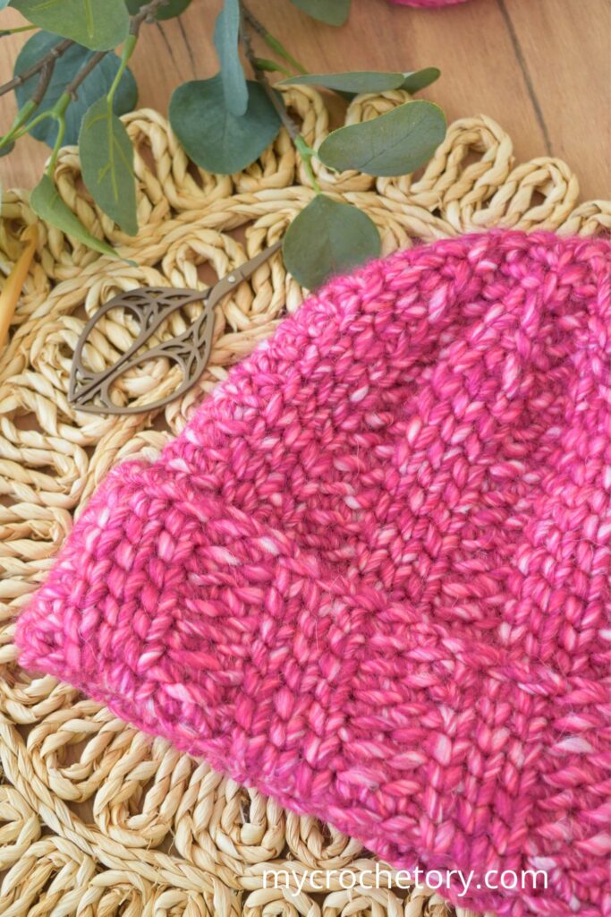 Chunky crochet hat in pink yarn styled with craft tools and greenery.