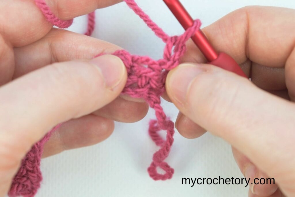 Inserting the hook into the vertical bars for the crochet waistcoat stitch, ensuring proper placement for a smooth and knit-like texture.