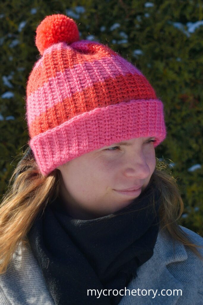 A cozy fast crochet hat pattern featuring bold pink and red stripes with a fluffy pom-pom.