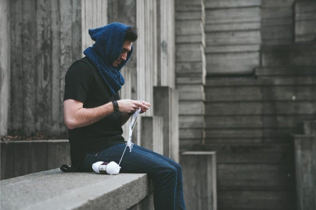 Man wearing a handmade blue crochet hooded scarf, sitting outdoors while crocheting with white yarn.