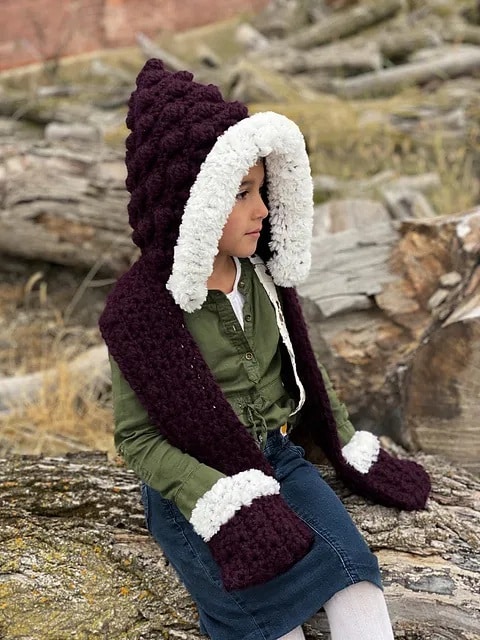Child wearing a chunky crochet hooded scarf with white trim and built-in pockets, sitting on wooden logs.