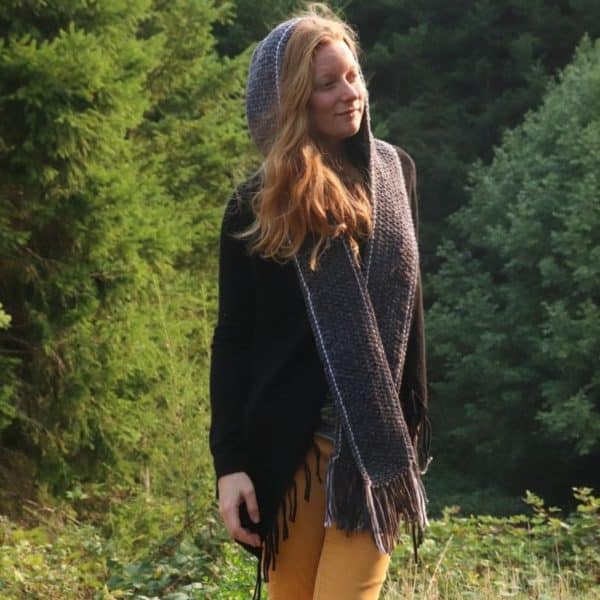 Woman standing outdoors wearing a gray crochet scarf with hood and fringe, made with soft yarn and simple stitches.