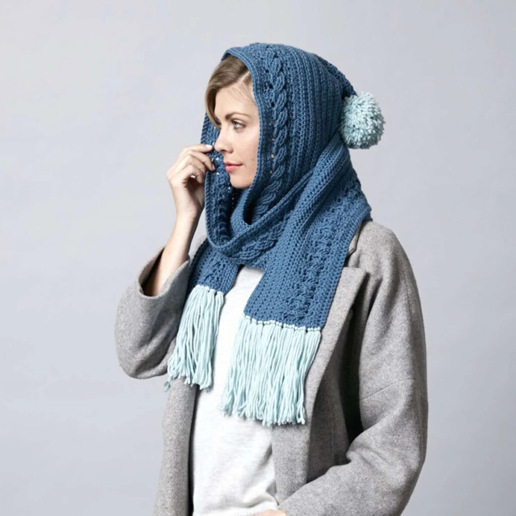 Woman wearing a blue cable crochet hooded scarf with light fringe and pompom detail, made with cozy winter yarn.