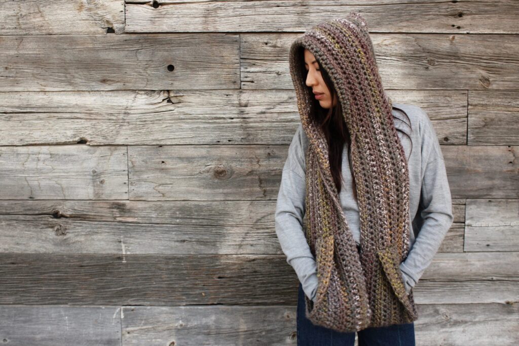Woman in a gray sweater wearing a chunky crochet hooded scarf with pockets in rustic brown tones.