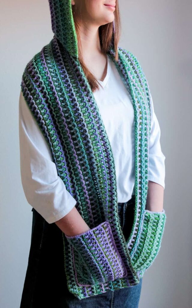 Crochet hooded scarf with pockets made in green, purple, and gray stripes using textured stitches.