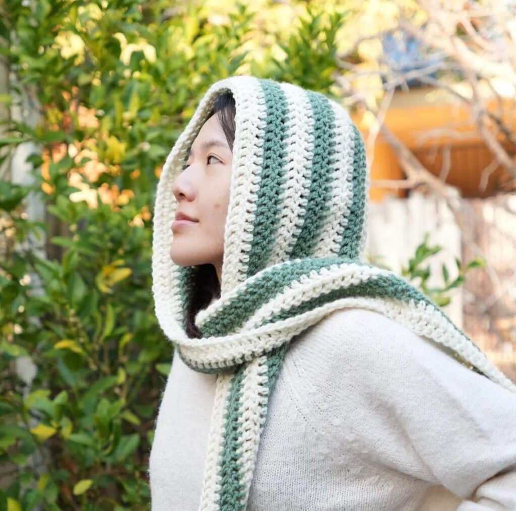 Woman wearing a green and white striped crochet hooded scarf, handmade with half double crochet stitches.