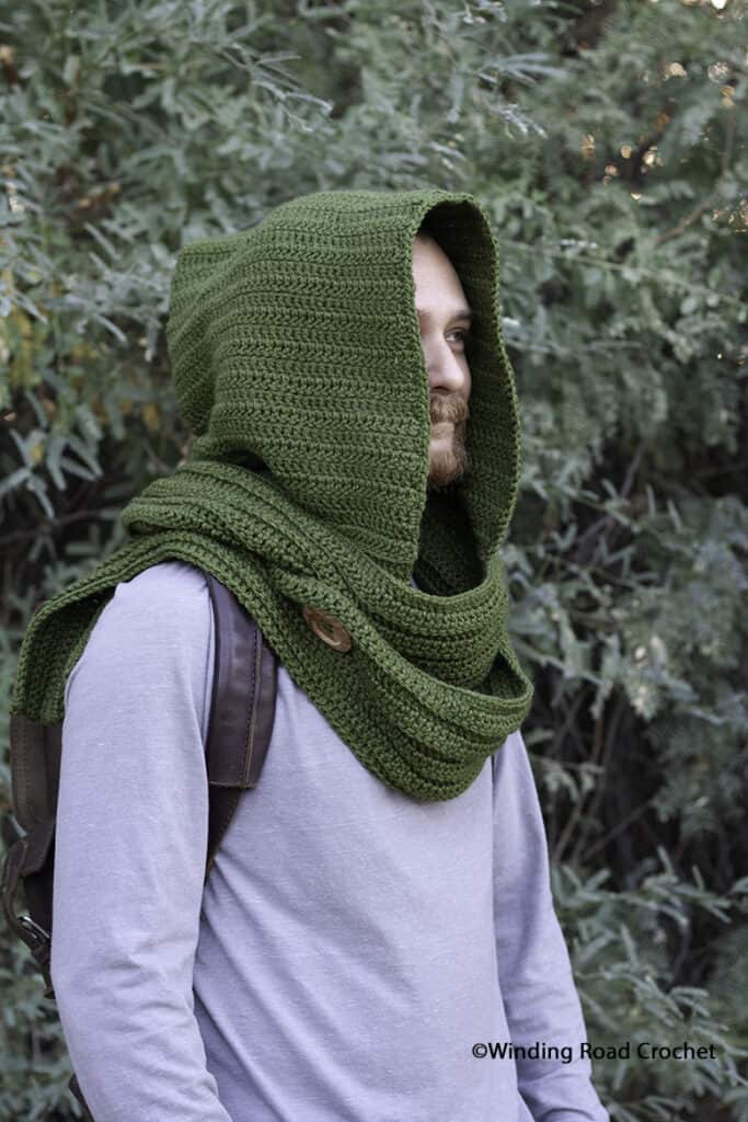 Man wearing a forest green crochet hooded scarf with button detail, cozy and rustic for outdoor adventures.