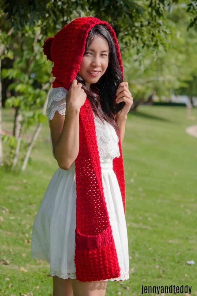 Woman outdoors wearing a bright red crochet hooded scarf with front pockets, inspired by Little Red Riding Hood.