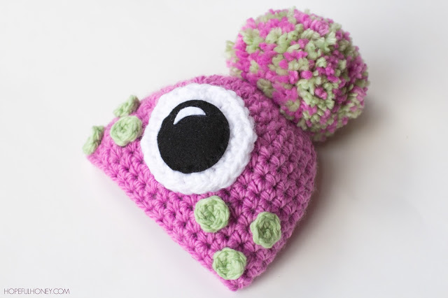 Pink crochet baby monster hat with one big eye, green freckles and a pom-pom