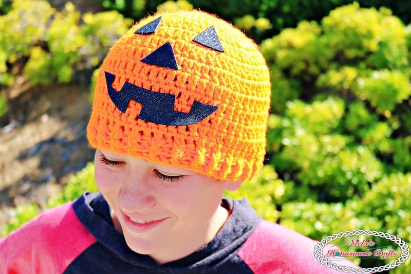 Orange crochet jack-o-lantern beanie with a black face worn by a smiling teen