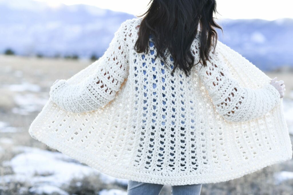 Back view of the Light Snow oversized crochet cardigan in cream with open eyelets, in a snowy field