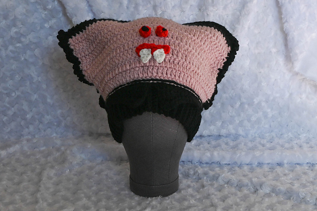 Pink crochet vampire sack hat with a black Dracula collar, bobble eyes and white fangs on a mannequin head
