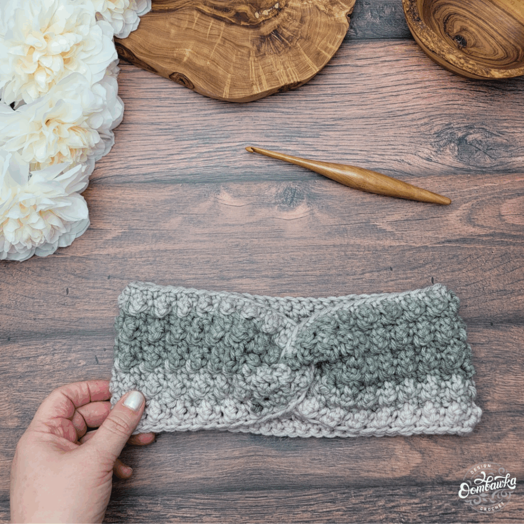 Textured twisted crochet ear warmer in sage green and gray on a wooden table with a crochet hook