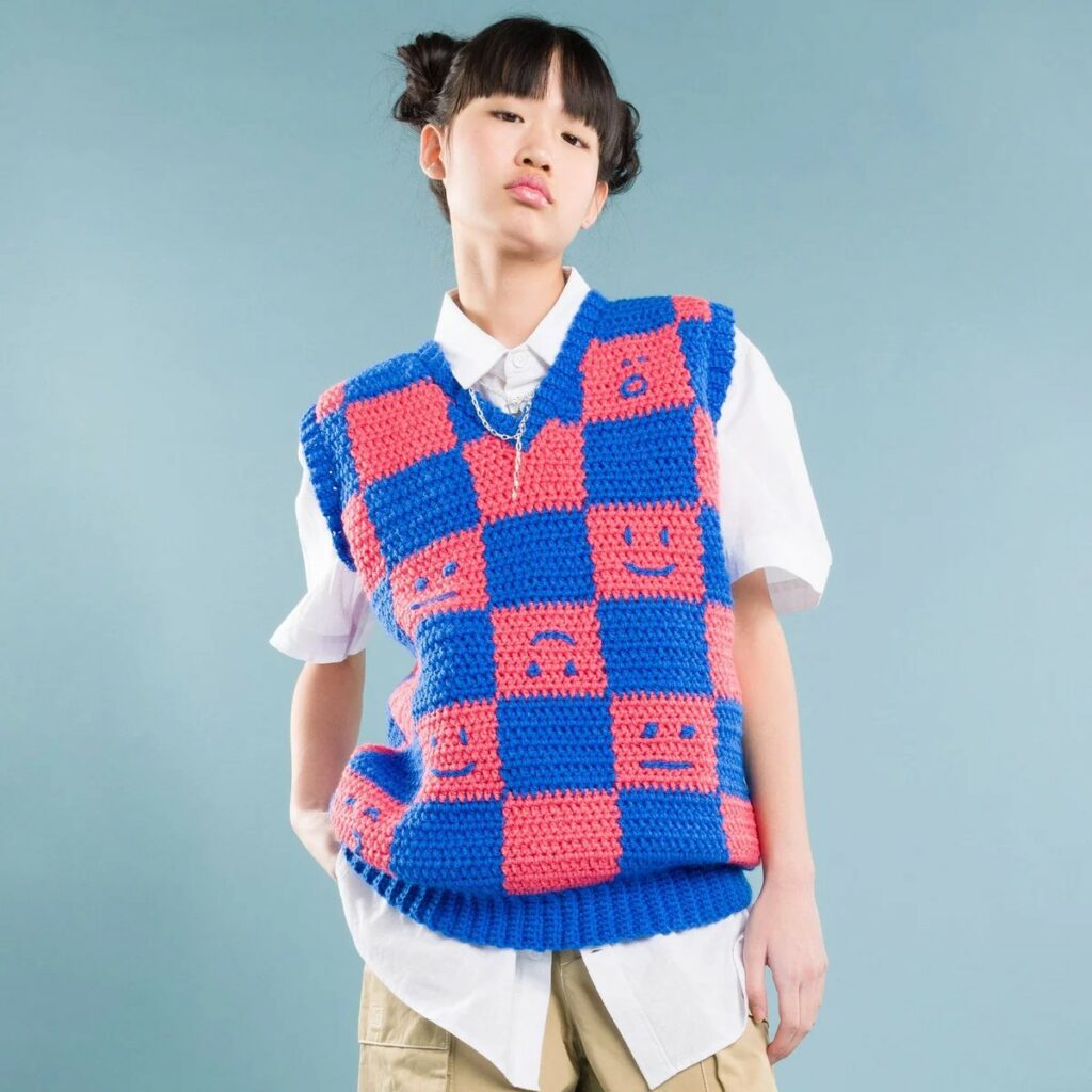 Crochet sweater vest with a blue and pink checkerboard pattern and smiley faces, worn over a white shirt