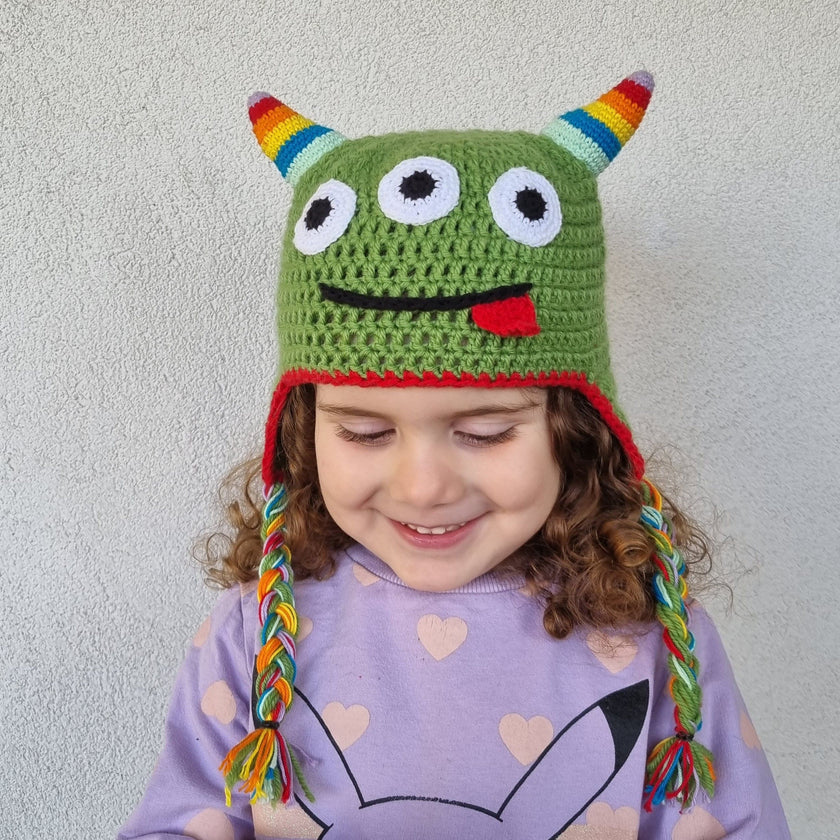 Green crochet monster hat with three eyes, rainbow horns and braided earflaps worn by a little girl