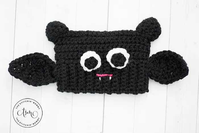 Black crochet baby bat sack hat with round ears, side wings, white eyes and tiny fangs, free Halloween hat pattern