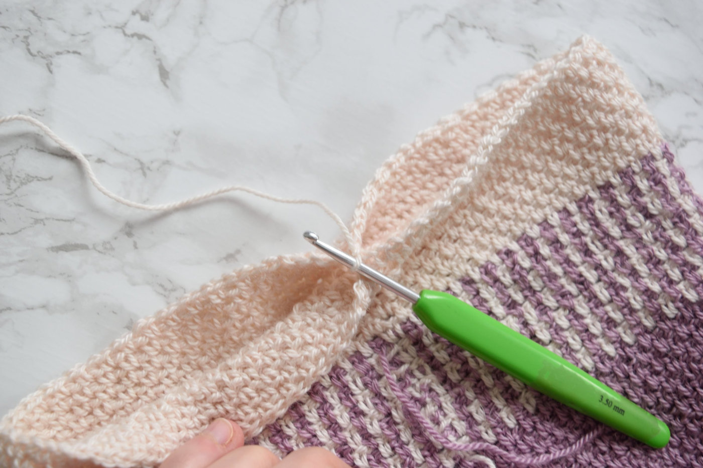 Closing the top of a purple and cream crochet beanie with a yarn tail and a 3.5 mm crochet hook on a marble table