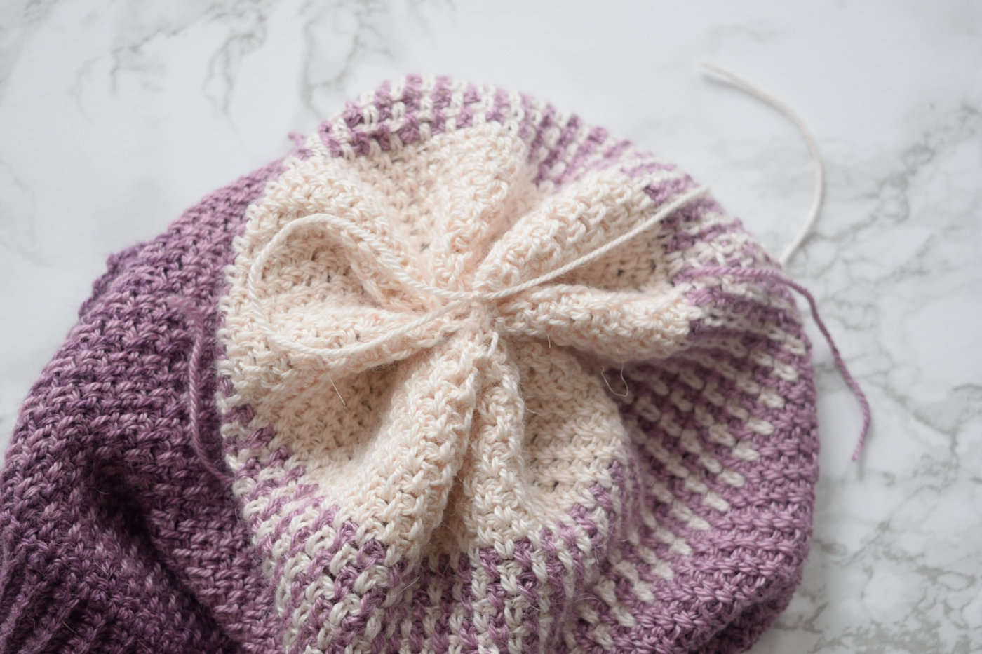 Gathered top of a purple and cream textured crochet beanie pulled closed with a yarn tail