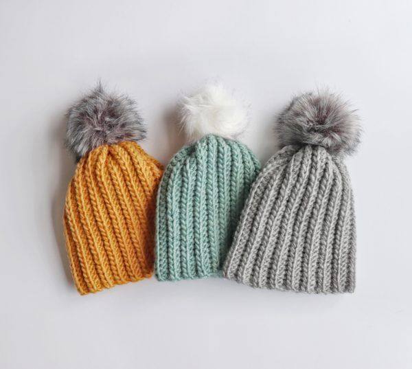 Three crochet winter hats in mustard, teal and gray with faux fur pom poms