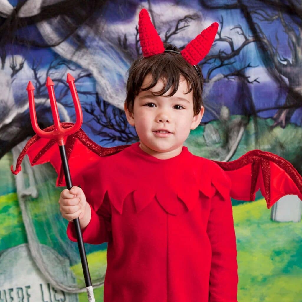 Little boy in a red devil costume with crochet devil horns on a headband, holding a pitchfork