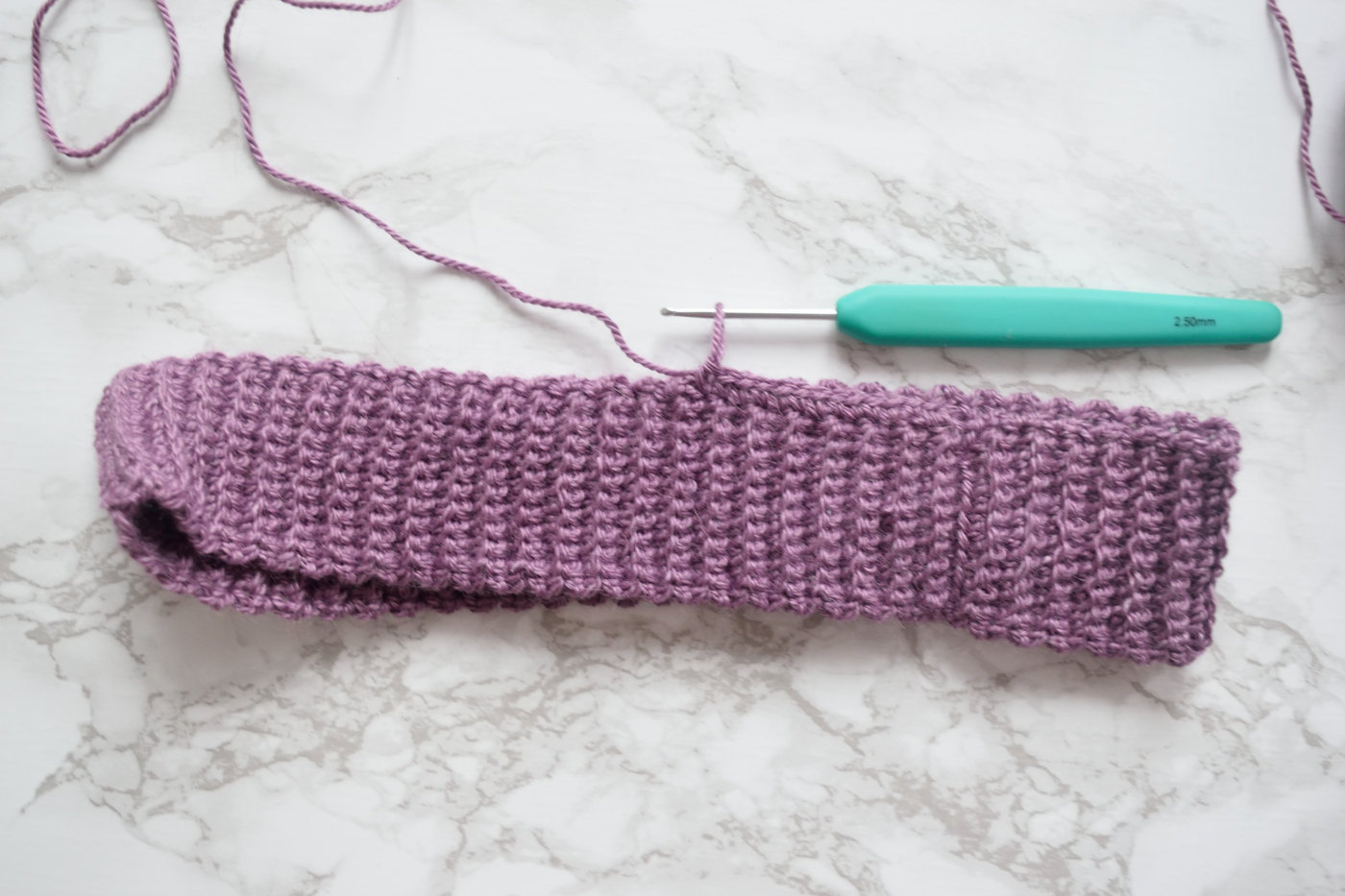 Purple crochet ribbing strip for a beanie brim next to a 2.5 mm crochet hook