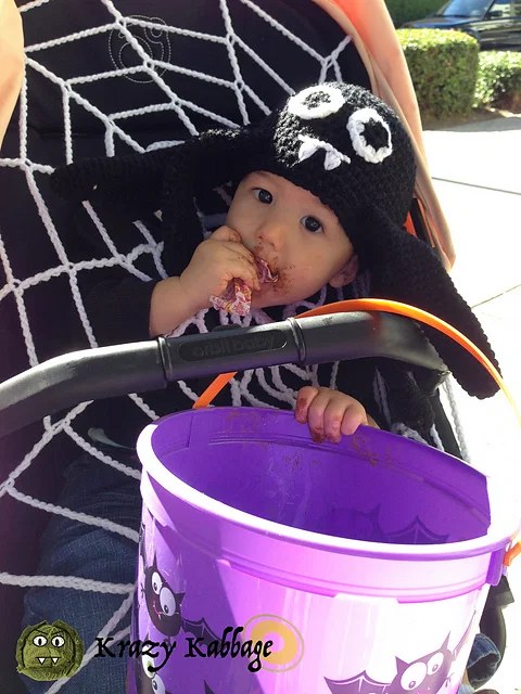 Toddler in a black crochet spider hat and white spider web poncho holding a trick-or-treat bucket