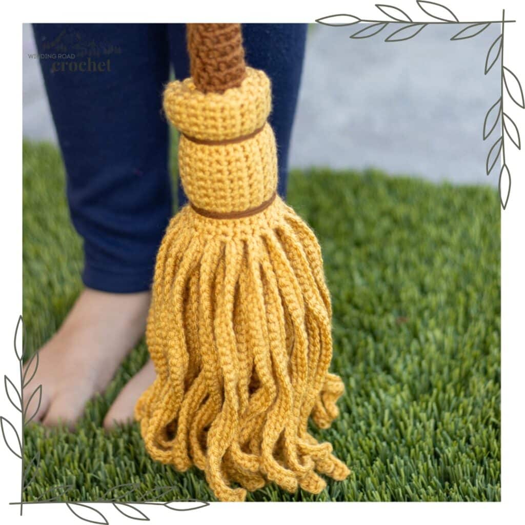 Yellow crochet witch's broom prop with a brown handle and yarn bristles on grass