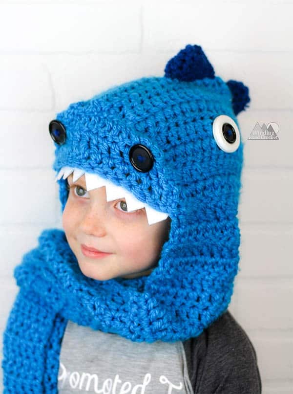 Child in a blue crochet dragon hood scarf with spikes, button eyes and white teeth