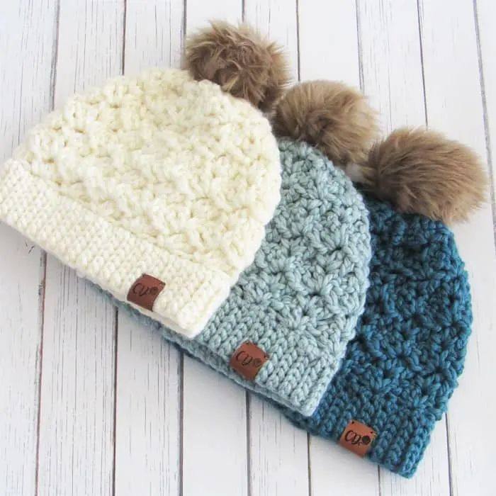 Three chunky crochet beanies in cream, light blue and teal with faux fur pom poms