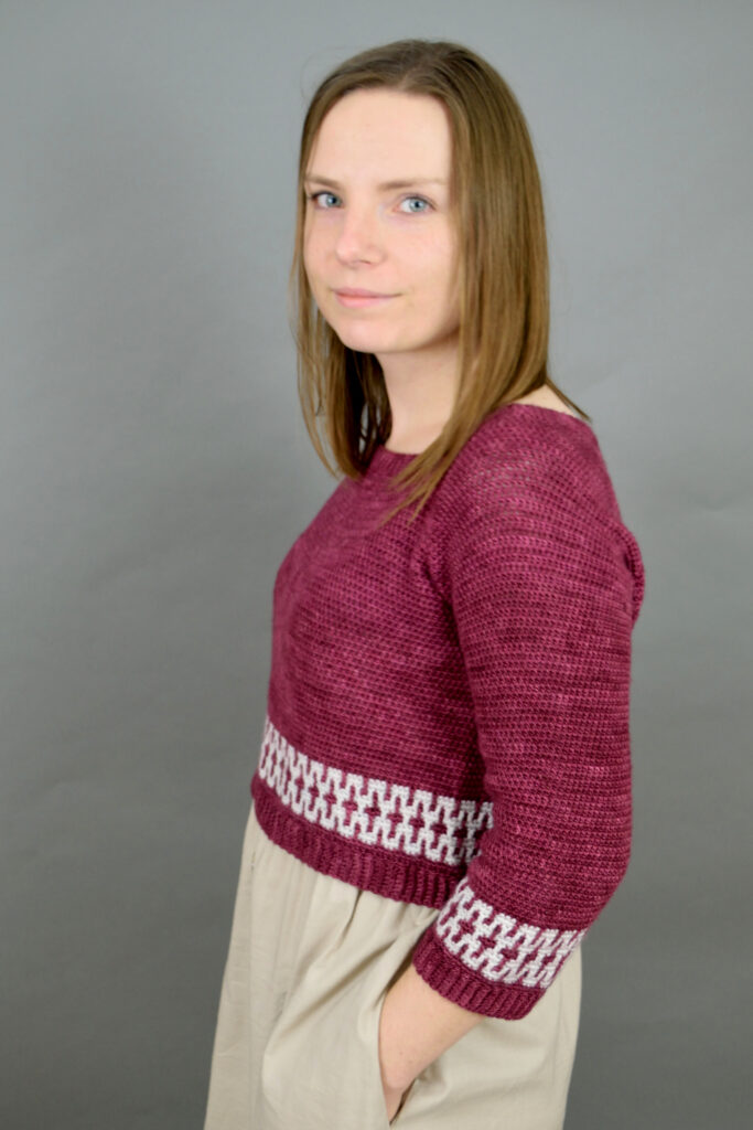 	Woman wearing a burgundy Mosaic Cropped crochet sweater with white mosaic bands at the hem and cuffs